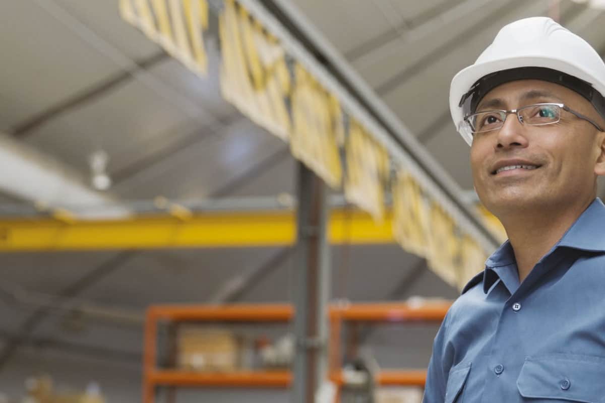Ingeniero usando casco de seguridad blanco y camisa de trabajo azul parado en planta de manufactura con equipo industrial en el fondo